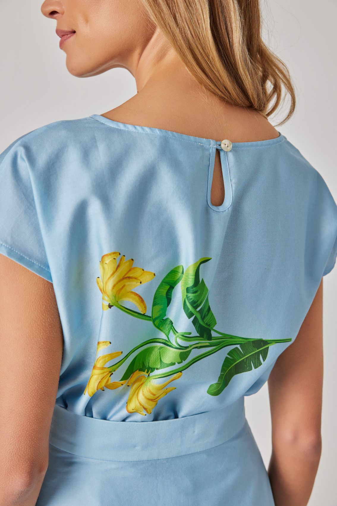 Model trägt hellblaues Kleid mit gelbem Blütenprint und Taillenbund, Rückenansicht. Tropfenausschnitt mit kleinem Knopfverschluss am hellblauen Kleid.