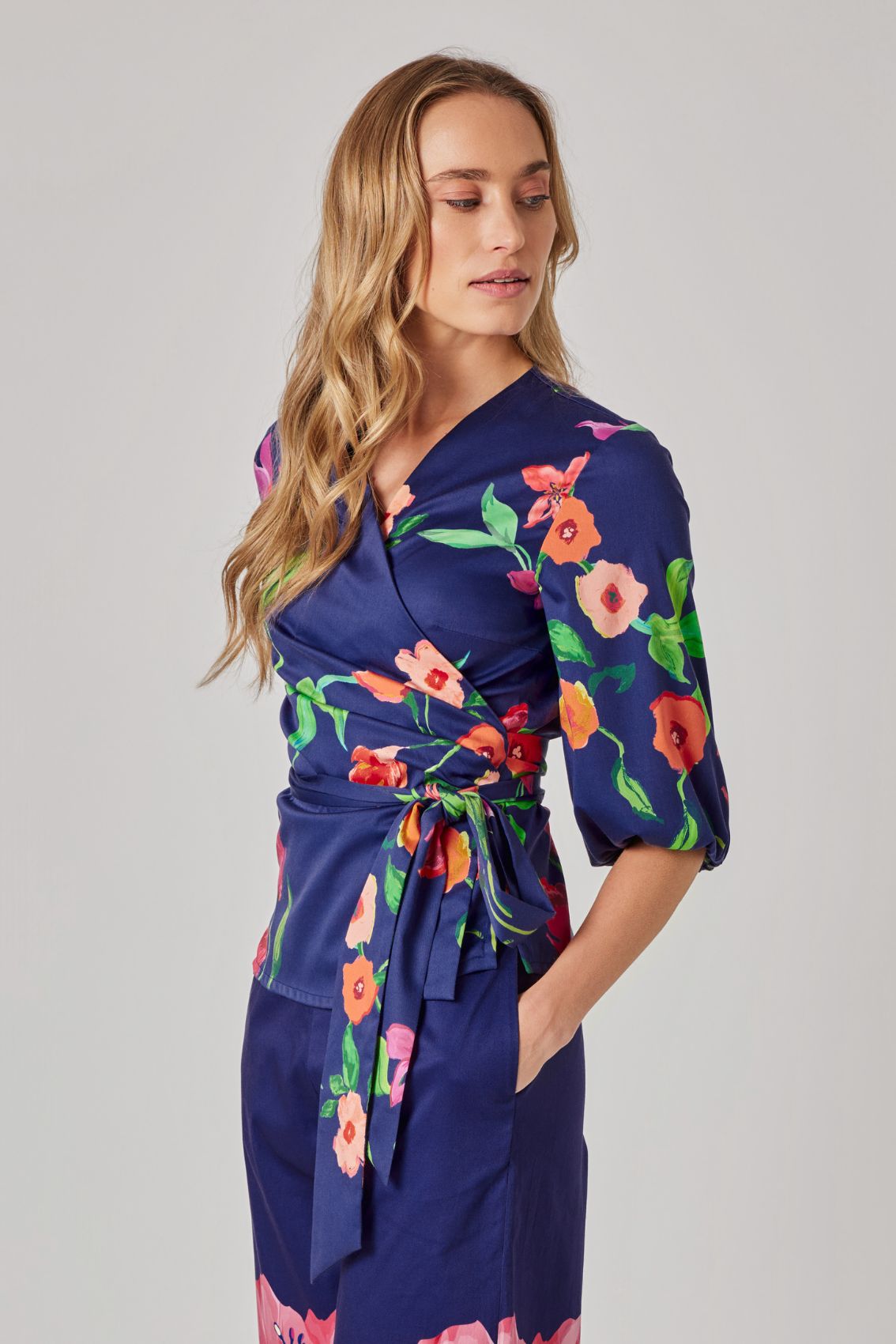 Model trägt Wickeloberteil in Dunkelblau mit floralem Print, V-Ausschnitt und Bindegürtel, Seitenansicht.