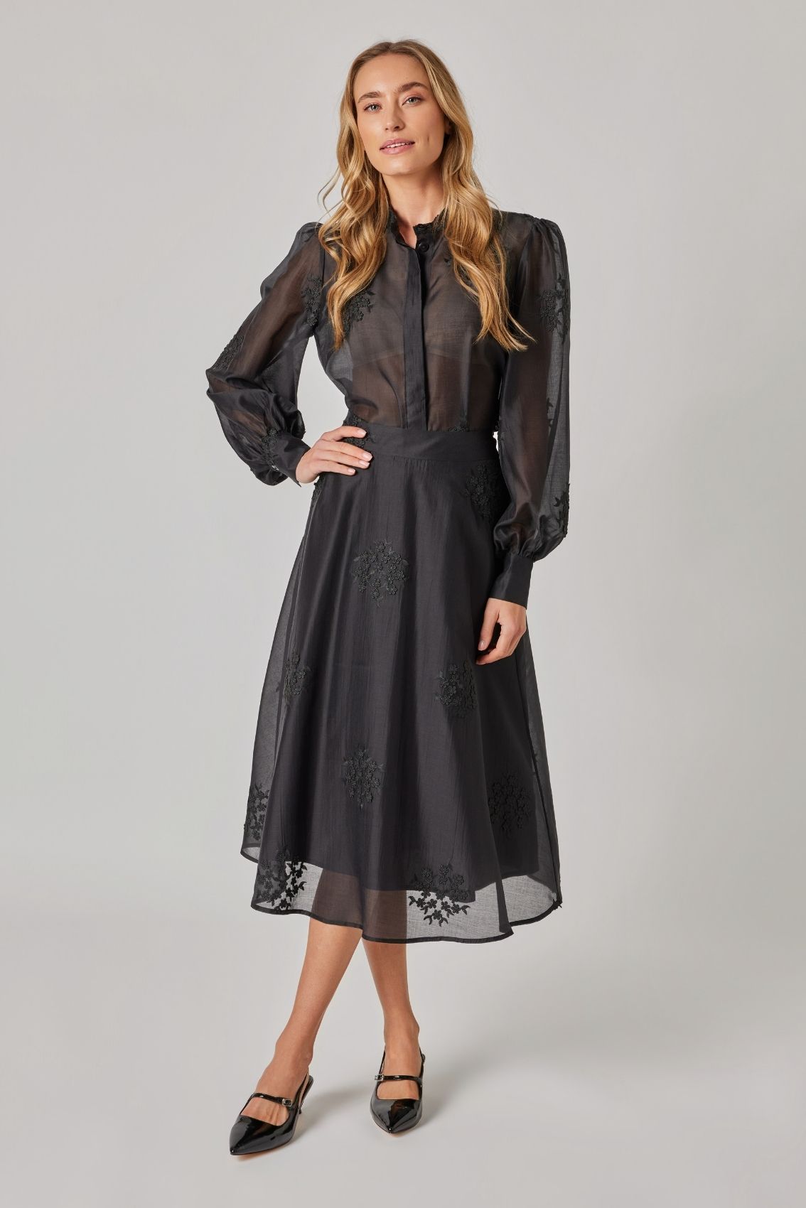 Model trägt schwarzen Midi-Rock mit floraler Stickerei und transparentem Oberstoff, Frontansicht.