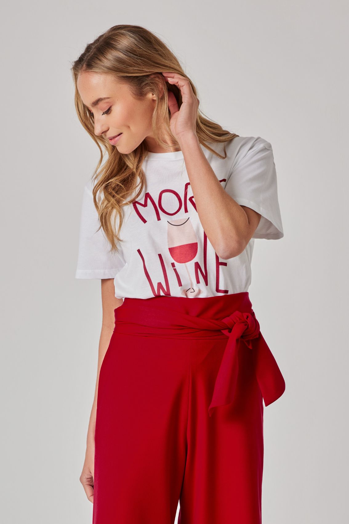 Model trägt weißes T-Shirt mit „MORE WINE“-Print; rote High-Waist Hose mit Bindegürtel