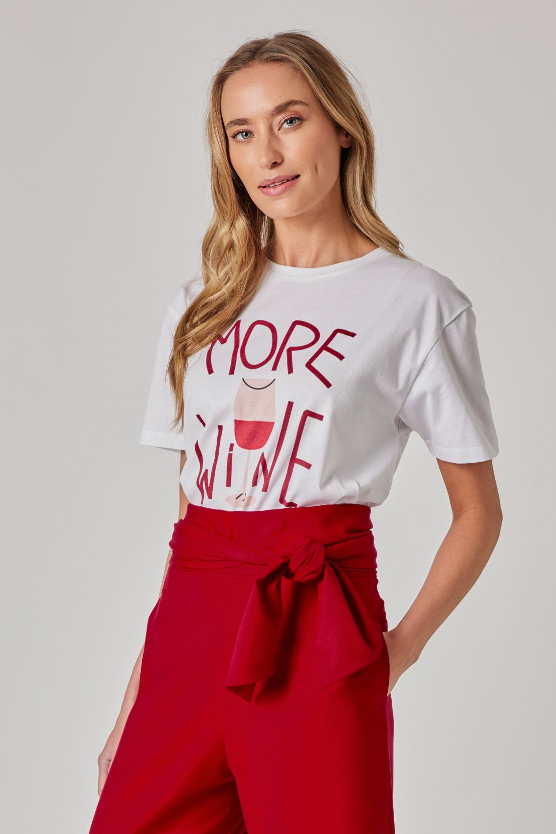 Model trägt weißes T-Shirt mit „MORE WINE“-Print; rote High-Waist Hose mit seitlicher Schleife