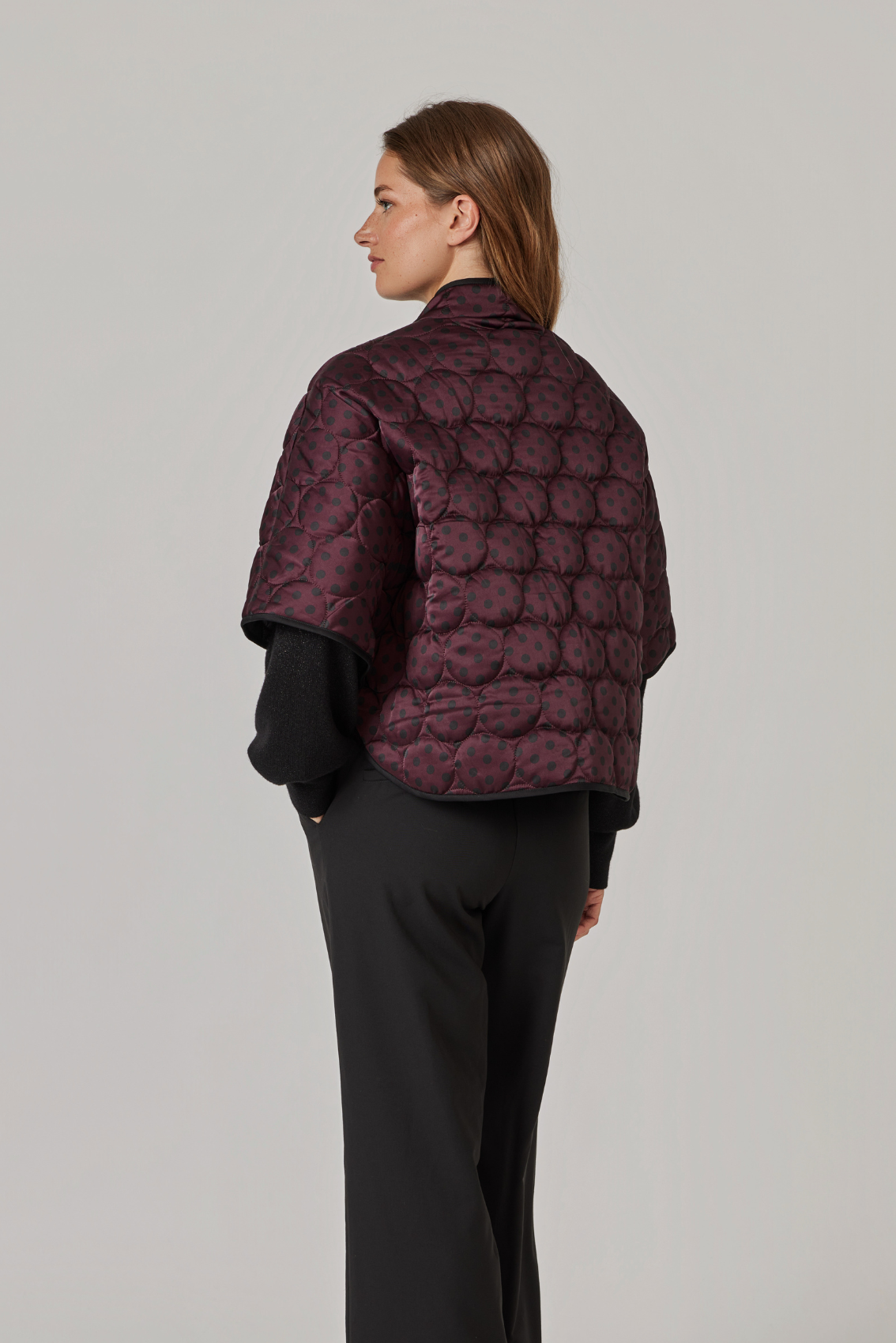 Model trägt Cape in Bordeaux mit Kreissteppung, Rückansicht mit schwarzer Paspel am Saum.