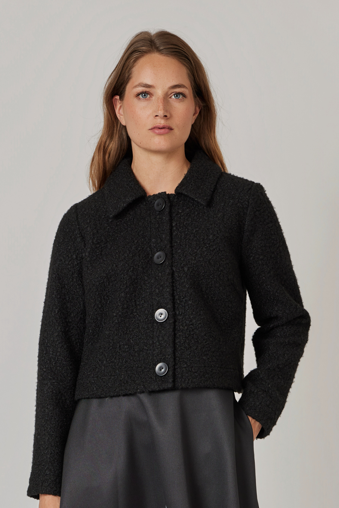 Model trägt kurze Bouclé-Jacke in Schwarz mit Hemdkragen zu dunklem Satinrock, Frontansicht.