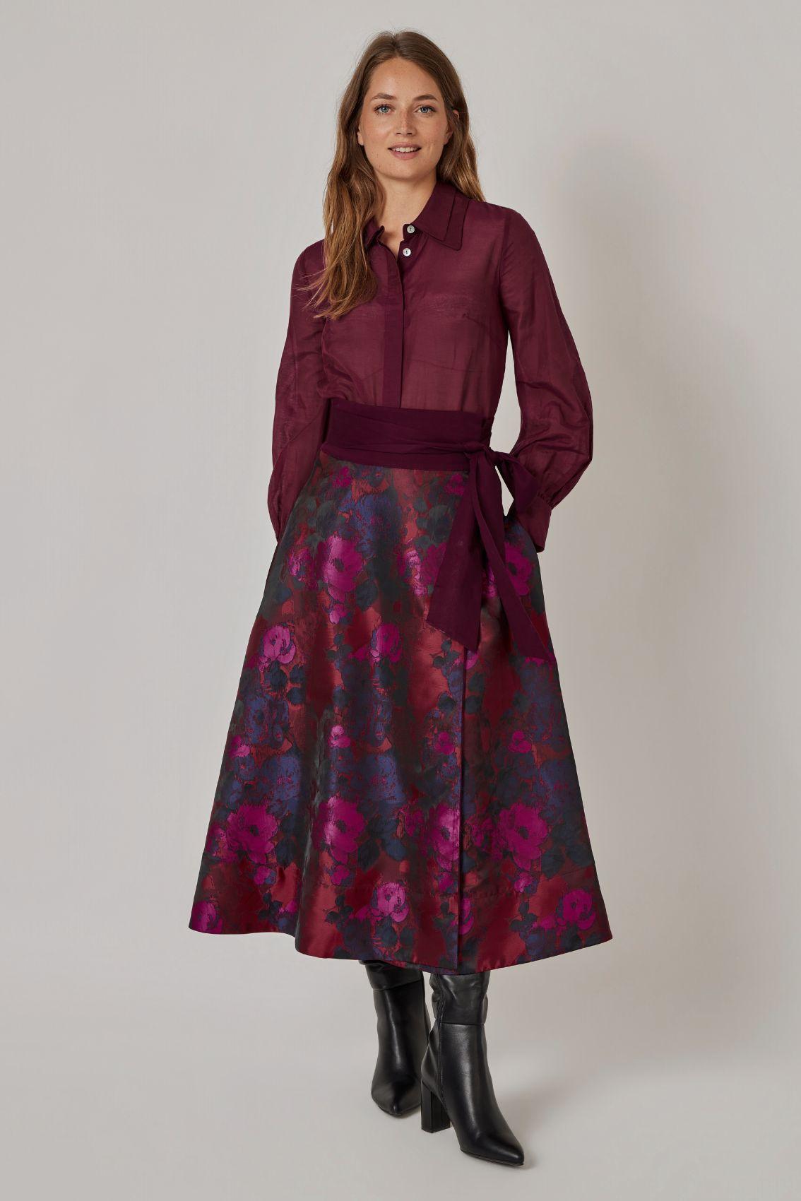 Model trägt Wickelrock mit Rosen-Jacquard, Bluse in Aubergine und schwarze Stiefel, Hand in der Seitentasche, Frontansicht.