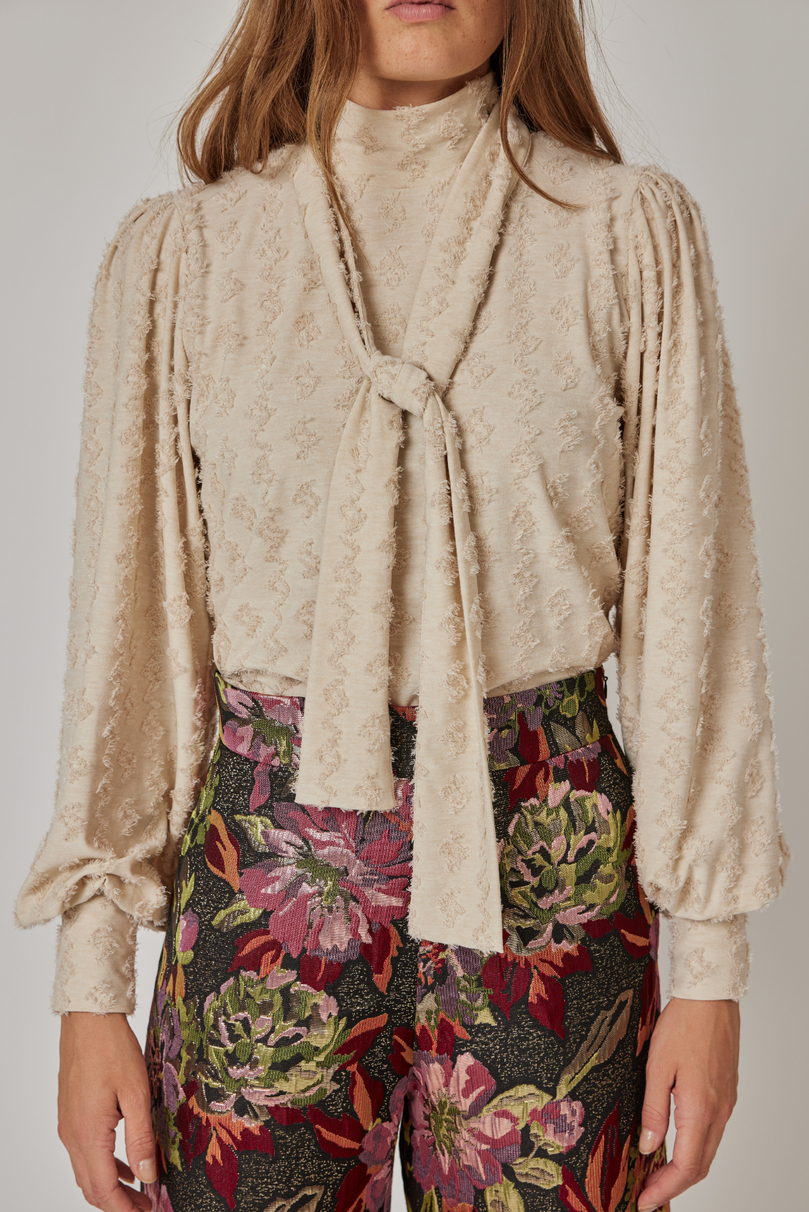 Model trägt Rollkragenshirt mit Schluppe in Creme, Schluppe vorne geknotet mit langen Enden, Frontansicht.
