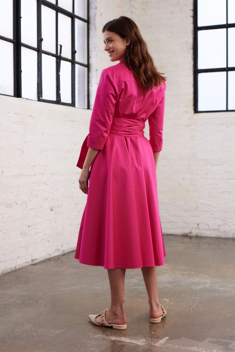 Modell trägt ein Blusenkleid mit Bindegürtel in einem leuchtenden Pink in Rückenansicht