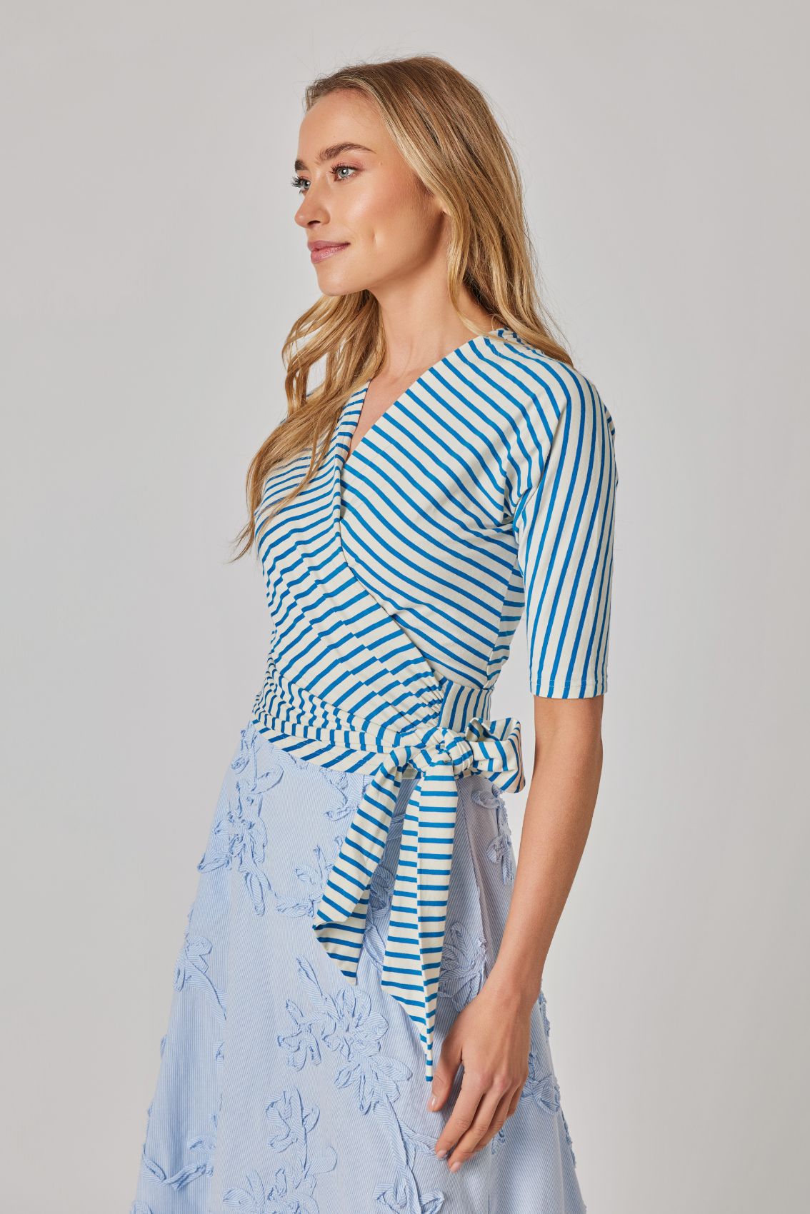 Model trägt Wickeltop blau-creme gestreift, Seitenansicht mit fest angenähter Bindung und 3/4-Ärmeln.