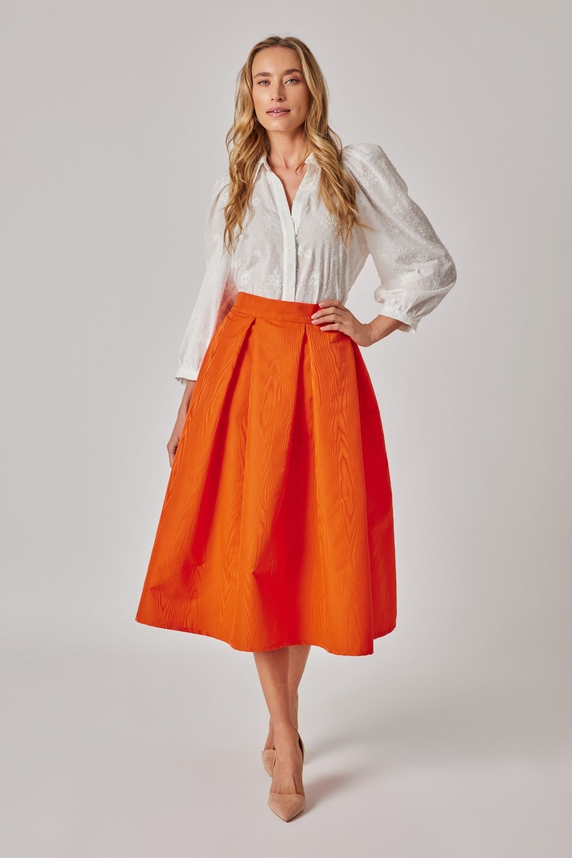 Model trägt Midirock in Orange mit Kellerfalten und Moiré-Effekt, Frontansicht mit taillenbetontem Sitz.