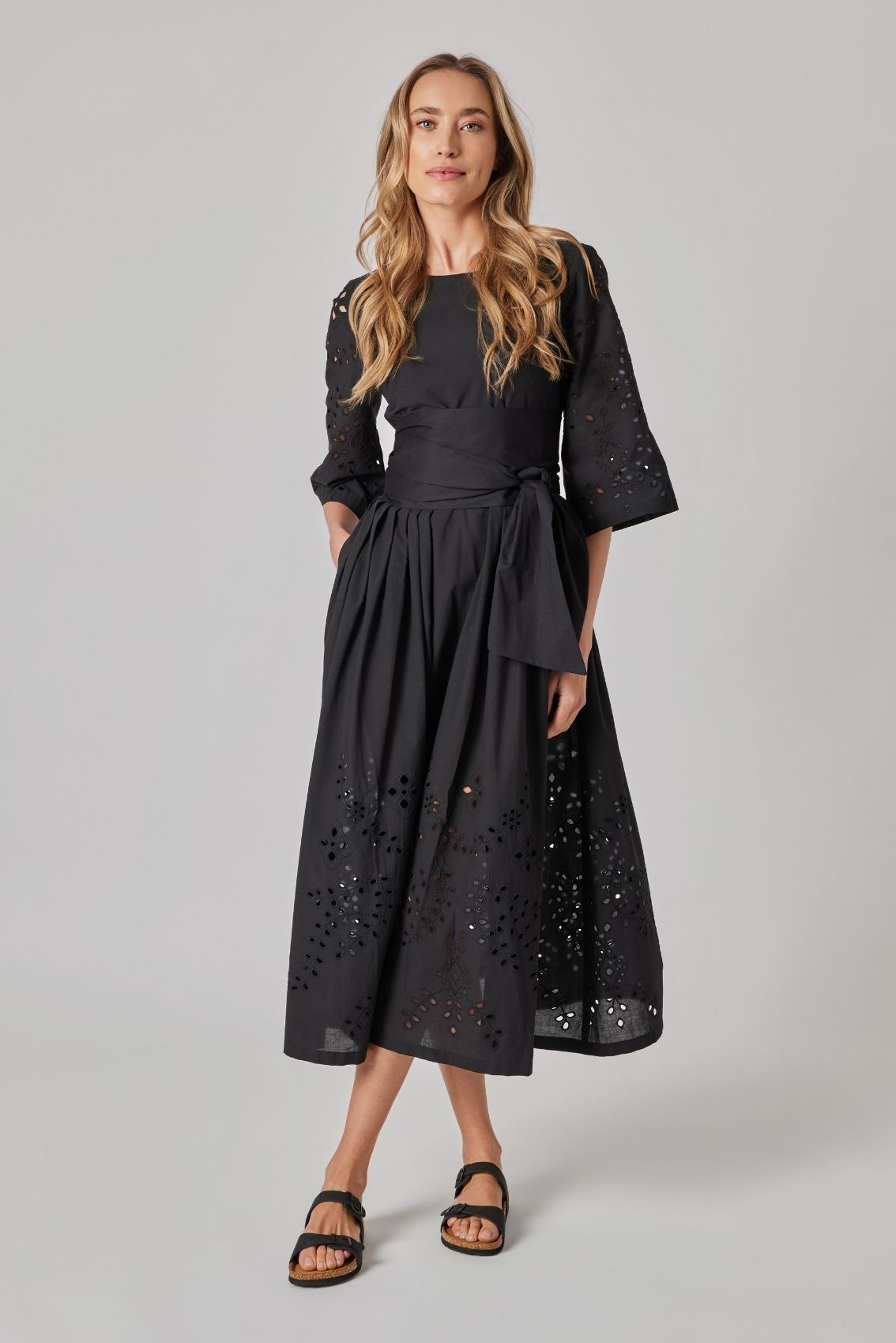 Model trägt schwarzes Midikleid aus Baumwolle mit Lochstickerei, Bindegürtel und Seitentaschen, Frontansicht.