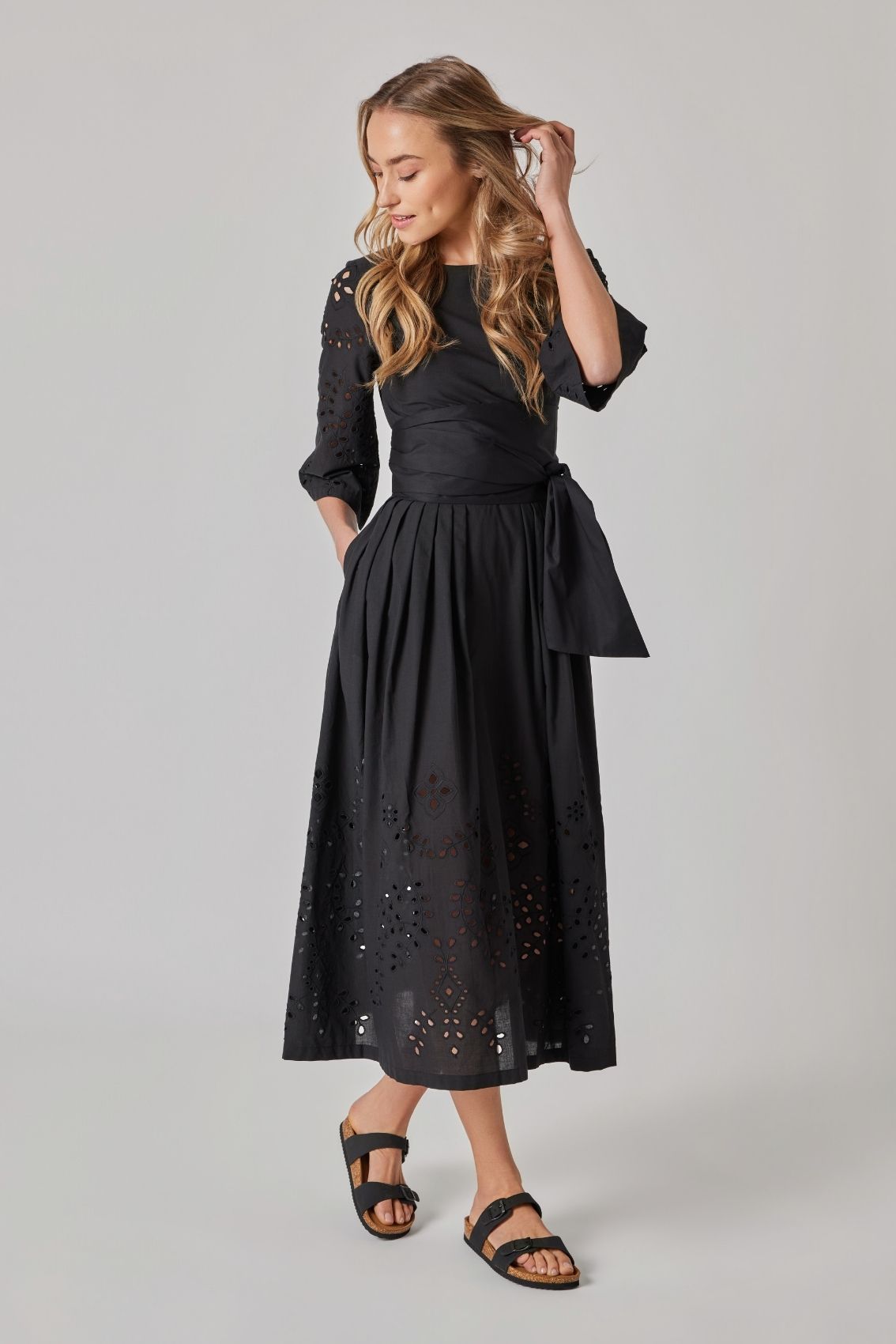 Model trägt schwarzes Midikleid mit tailliertem Bindegürtel und Lochstickerei an den Ärmeln, Seitenansicht.
