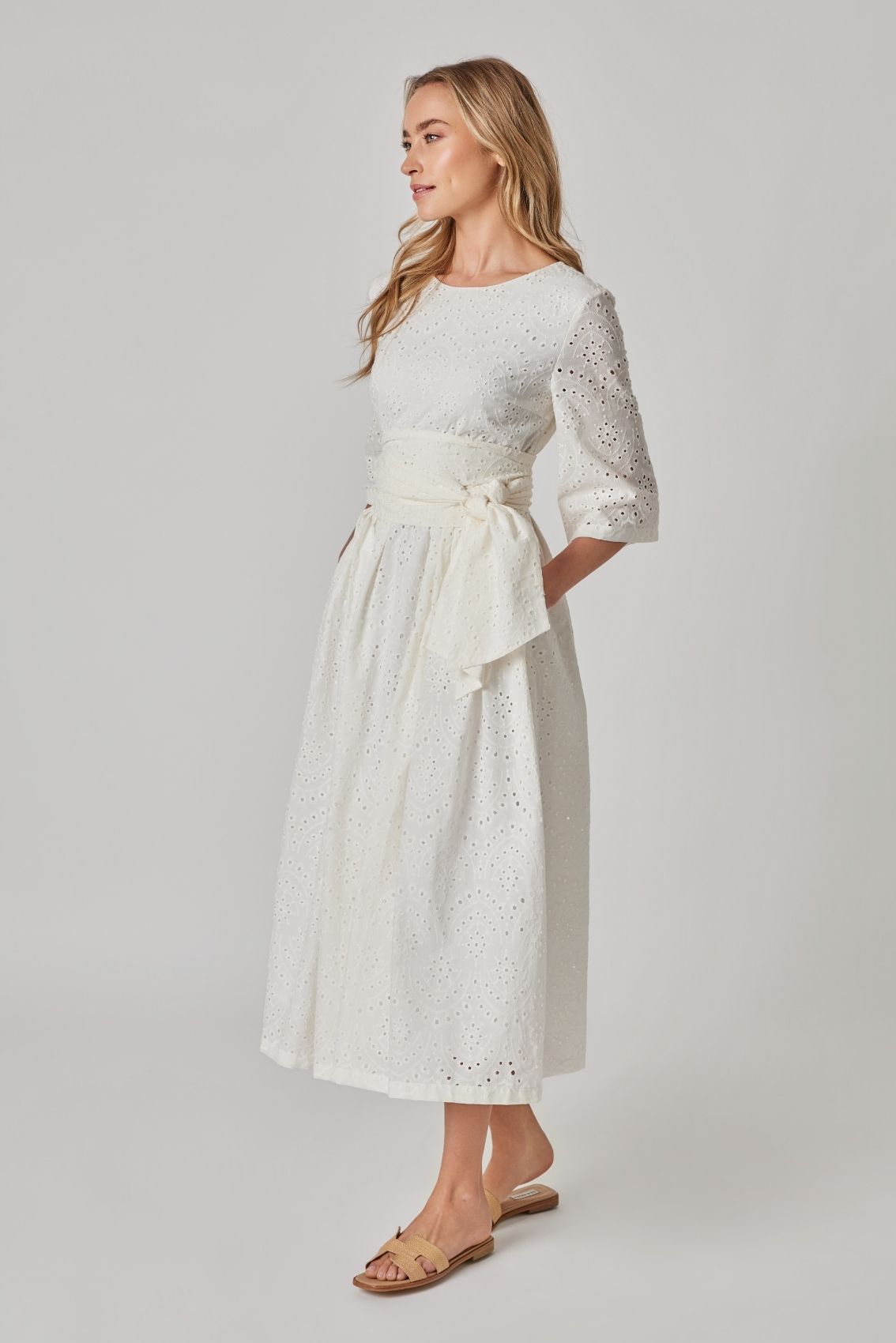 Model trägt weißes Broderie Midikleid mit taillierendem Bindegürtel und weitem Rock, Seitenansicht.