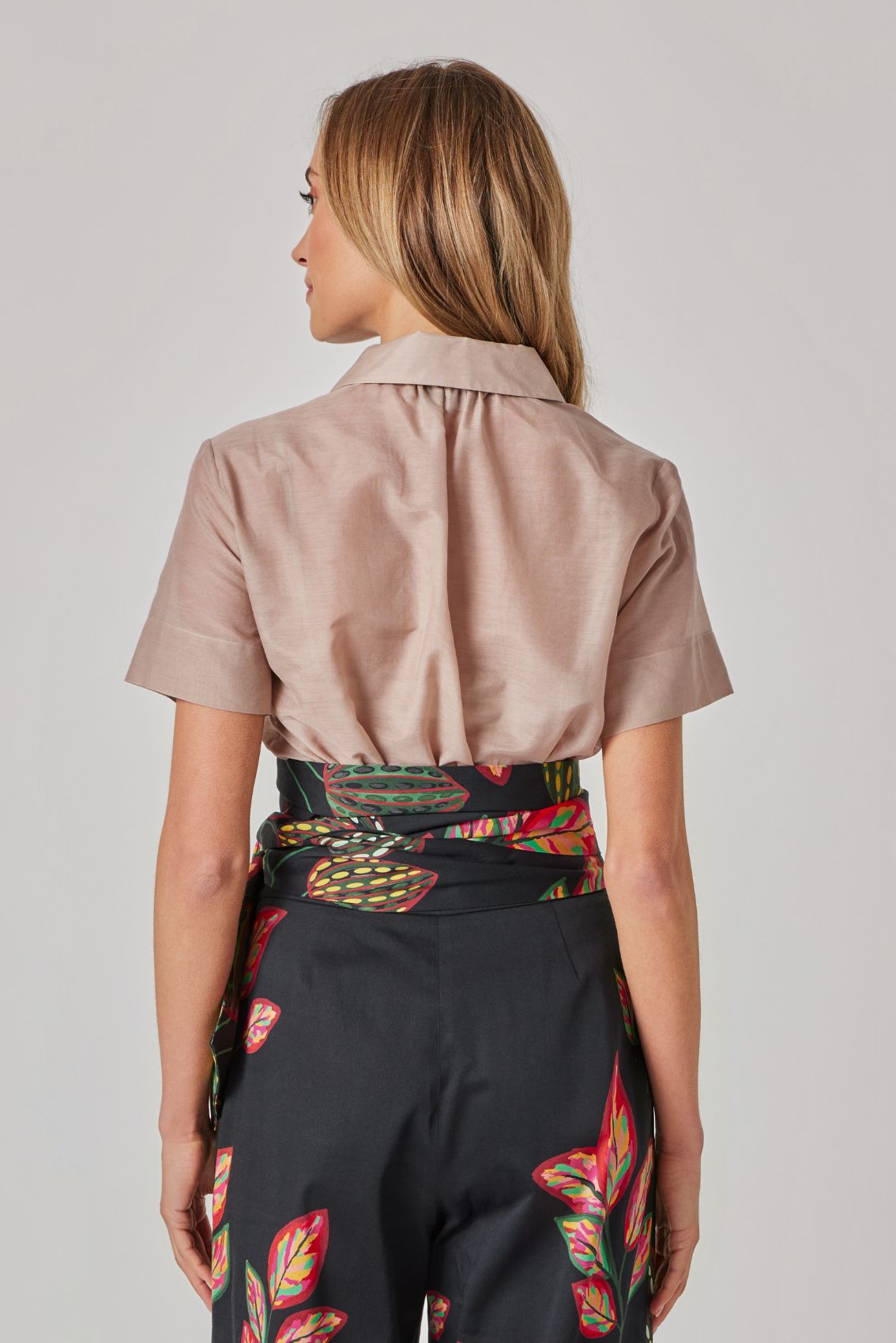 Model trägt beige, kurzärmelige Hemdbluse von hinten; hohe schwarze Hose mit Print und breitem Bindegürtel, Rückenansicht.