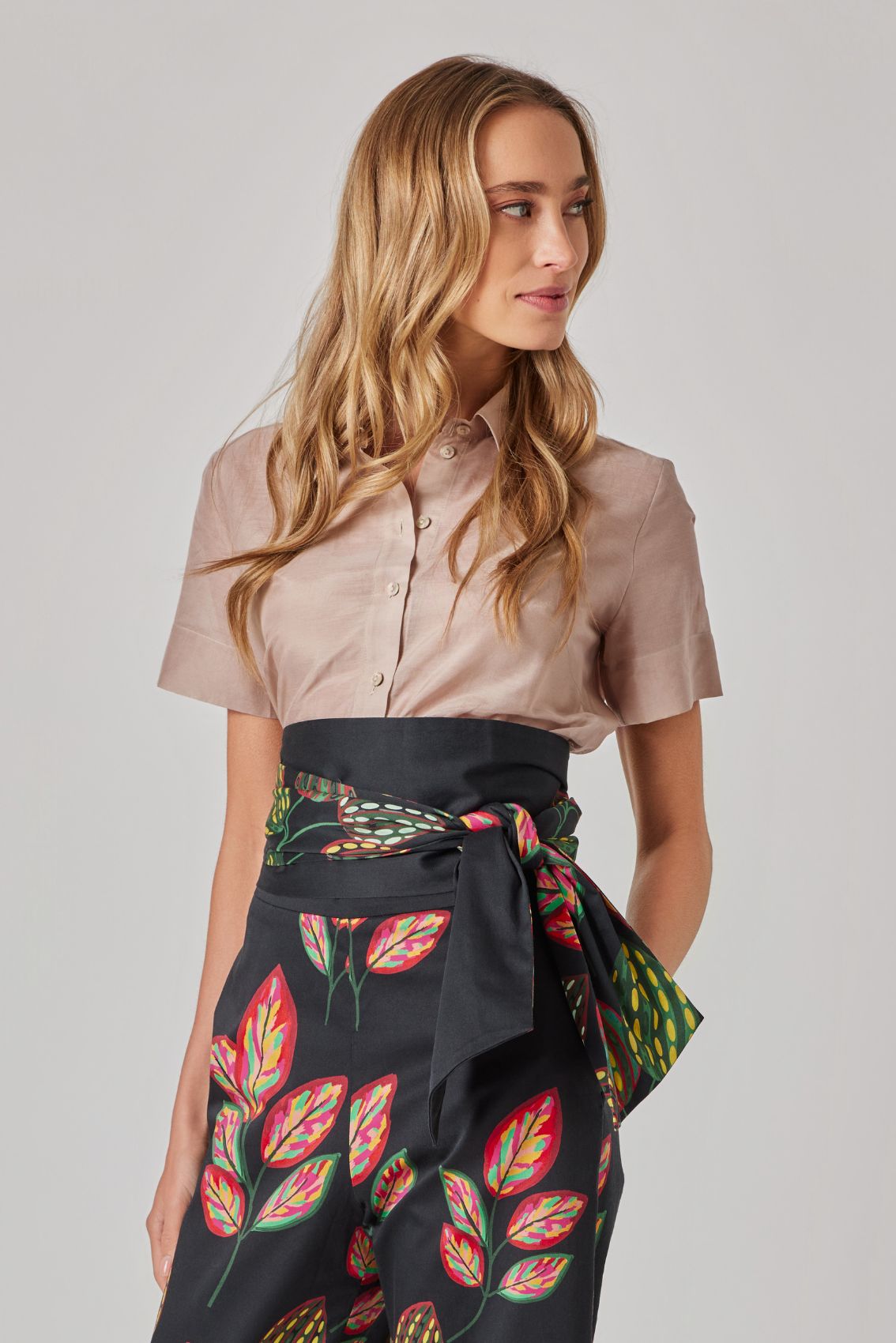 Model trägt beige, kurzärmelige Hemdbluse mit Kragen; hohe schwarze Hose mit Print und Bindegürtel, Seitenansicht.