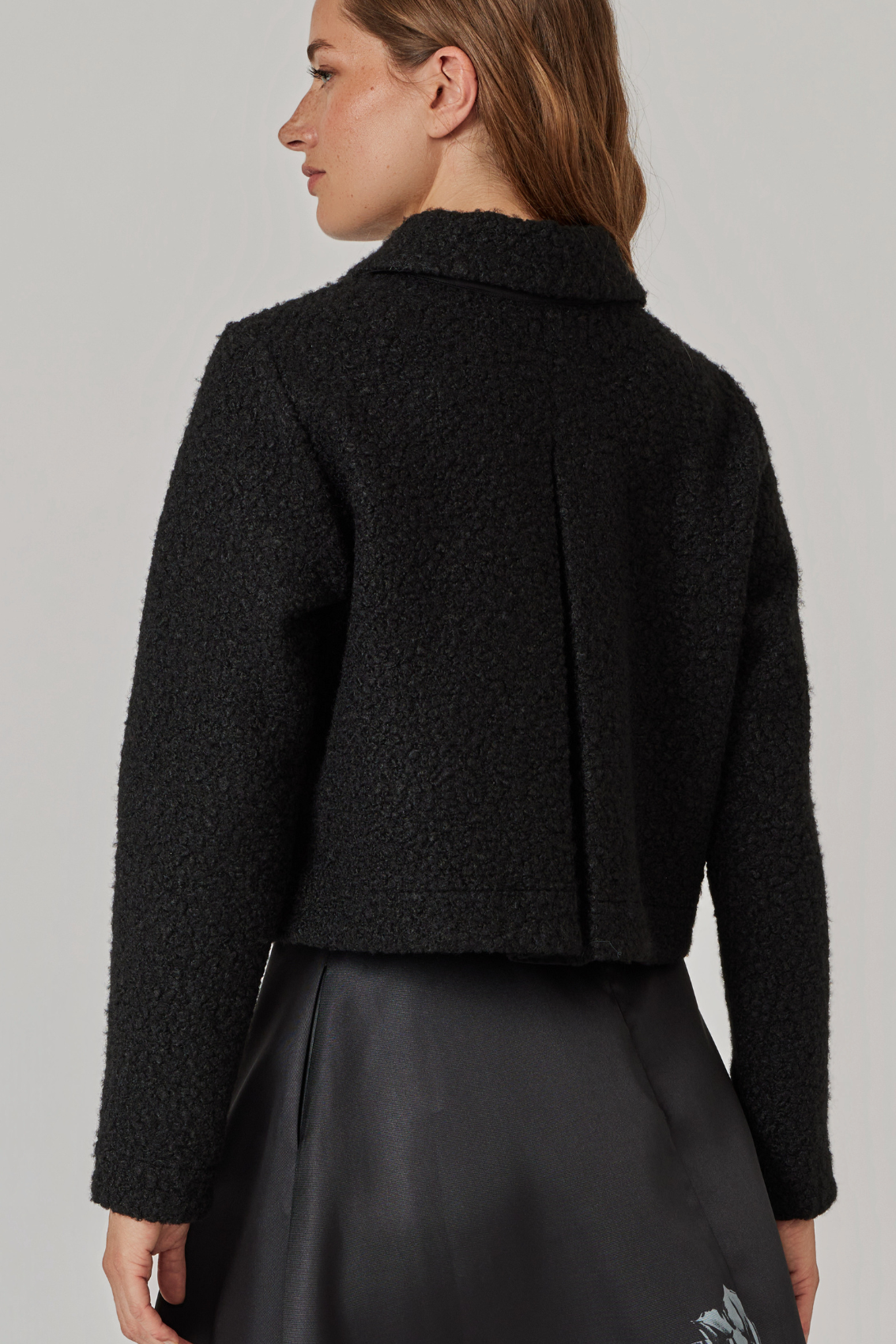 Model trägt schwarze Jacke, Rückenansicht mit mittiger Kellerfalte bis zum Saum.