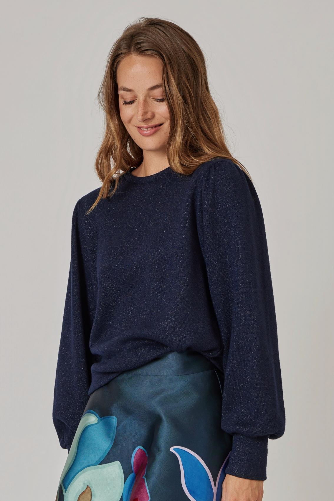 Model trägt navyfarbenen Pullover mit feinem Glitzer und gekräuselten Schultern, leicht seitliche Ansicht.