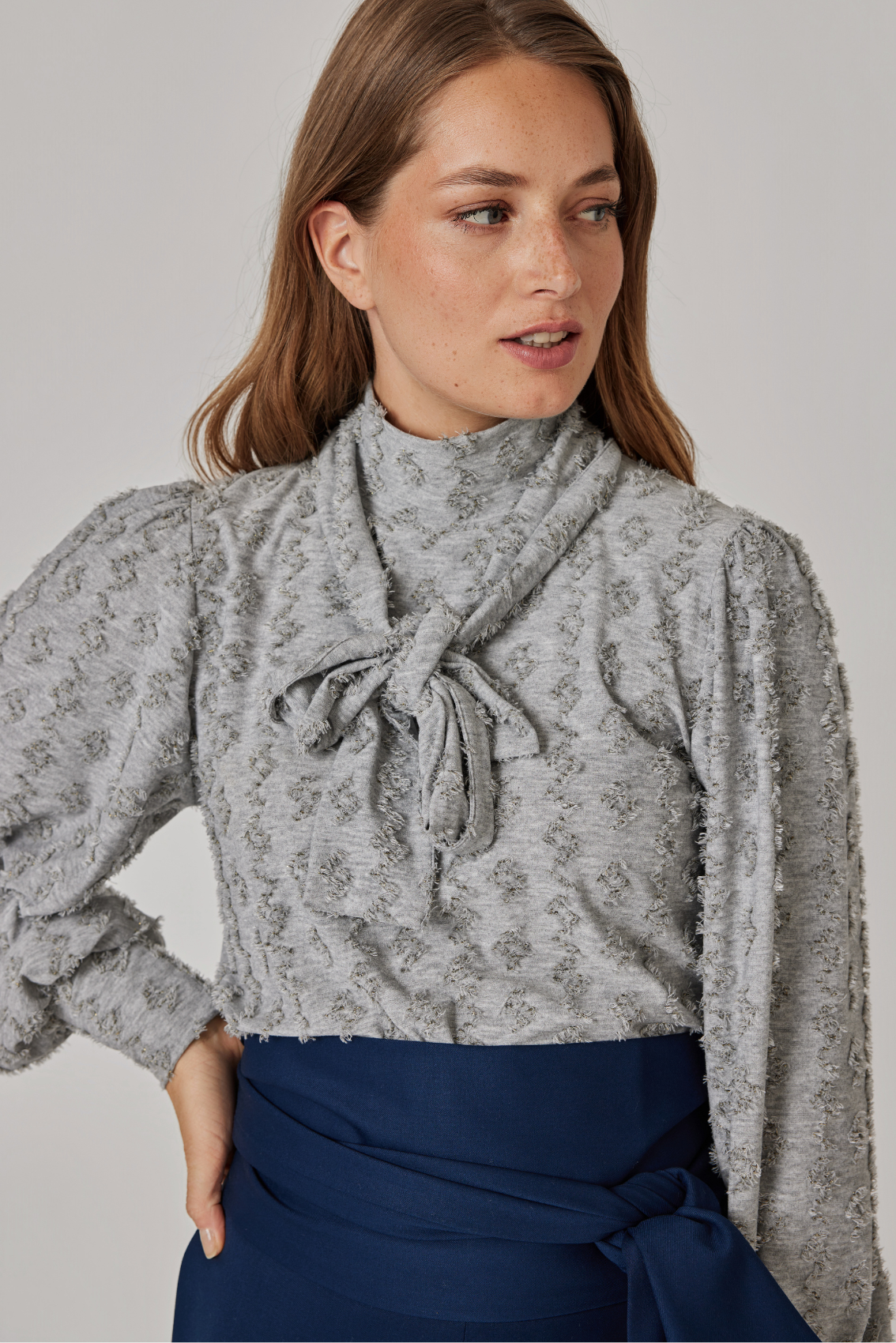 Model trägt Rollkragenshirt mit Schluppe in Steingrau, Schleife vorne gebunden, Ballonärmel, Detailansicht.