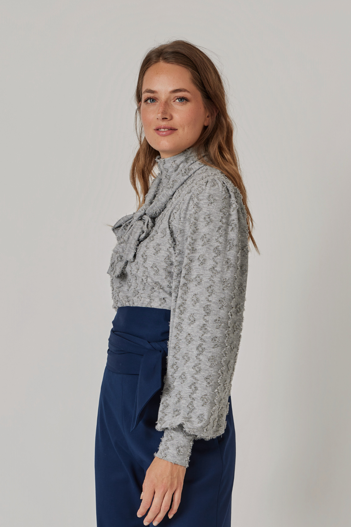 Model trägt steingraues Shirt mit gebundener Schluppe, Seitenansicht mit Ballonärmeln und breiten Manschetten.